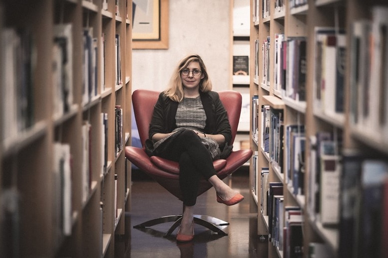 A woman (Heather Allansdottir) sits in an armchair with bookshelves on either side of her.