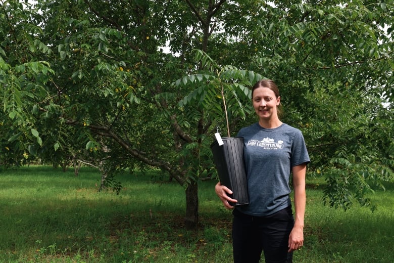 A woman stands holding a small sapling in front of a larger tree